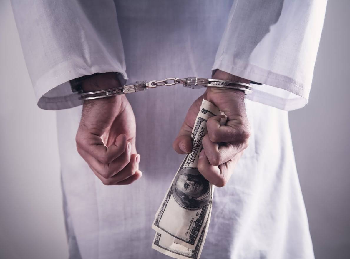 healthcare fraud; doctor in handcuffs while holding dollar bills